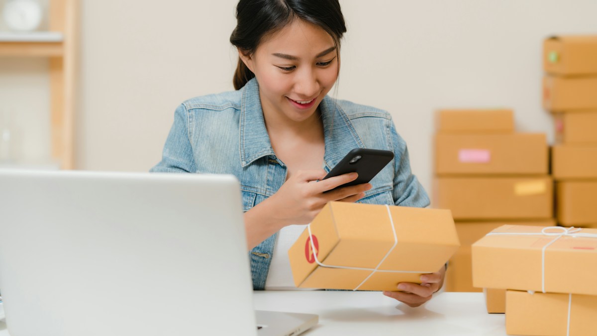 Une femme vérifie un colis sur son téléphone, près d’un ordinateur portable et de cartons